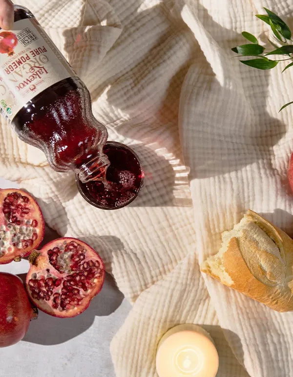 Bottle of pomegranate juice being poured onto a textured surface with pomegranate pieces and a lemon.