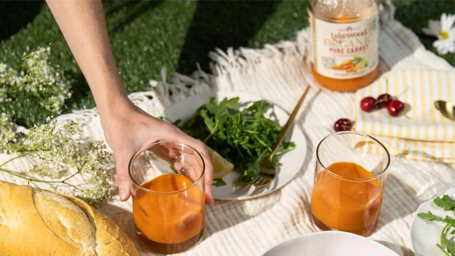 Person holding a glass of orange juice at a picnic with a bottle of juice and fruits on a blanket.