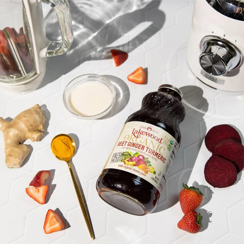 Bottle of Lakewood Organic Beet Ginger Turmeric on a white surface with ingredients like strawberries and ginger.